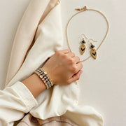 Bold statement bracelet, layered necklace, and earrings arranged on a neutral fabric backdrop with soft clothing accents.
