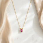 Load image into Gallery viewer, A delicate gold chain necklace with a rectangular emerald-cut pink gemstone pendant, displayed on a white marble surface framed by soft beige satin drapery.